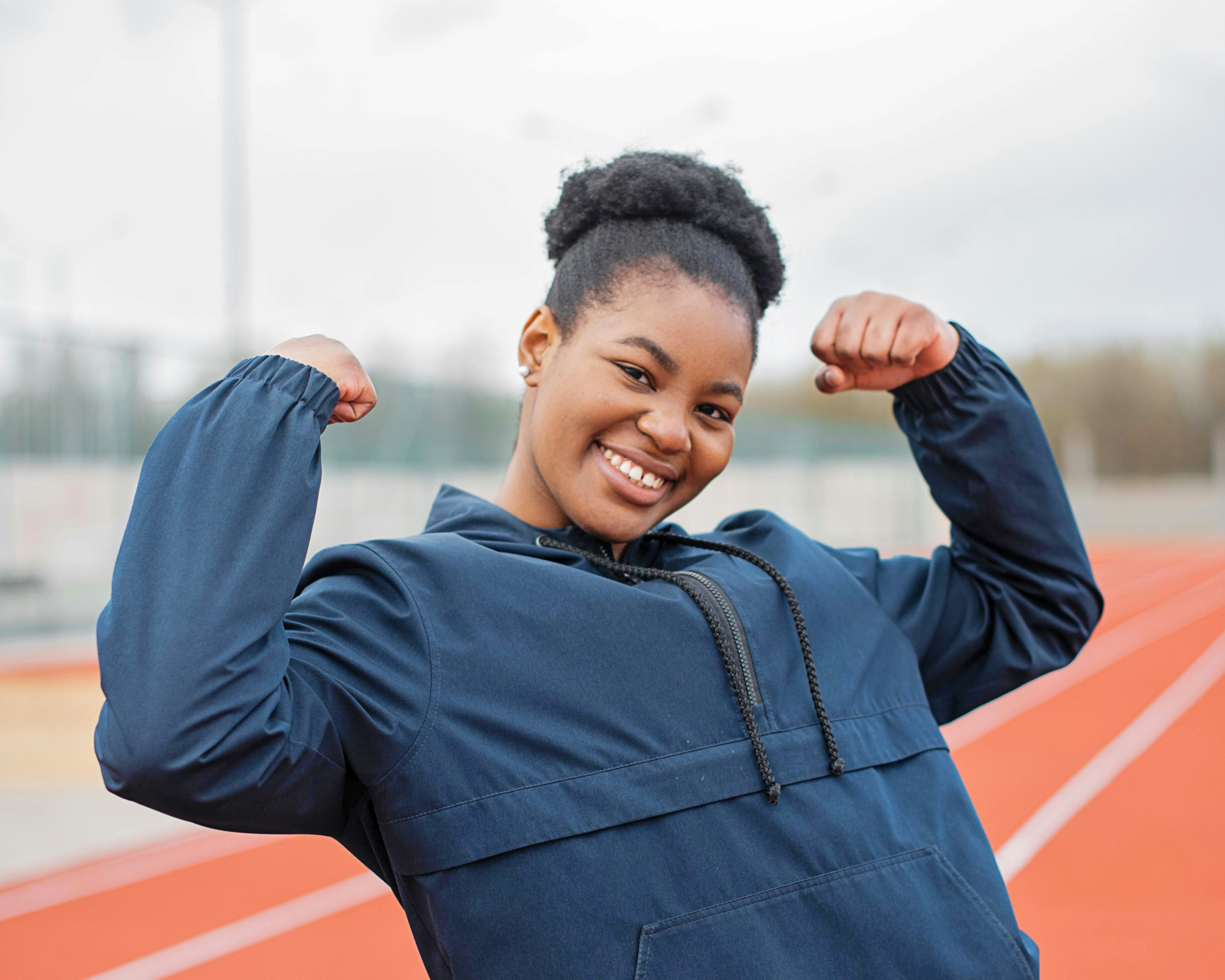 A smiling Black woman stands on a running track, flexing her arms in celebration of International Women’s Day 2025 and the #AccelerateAction theme.
