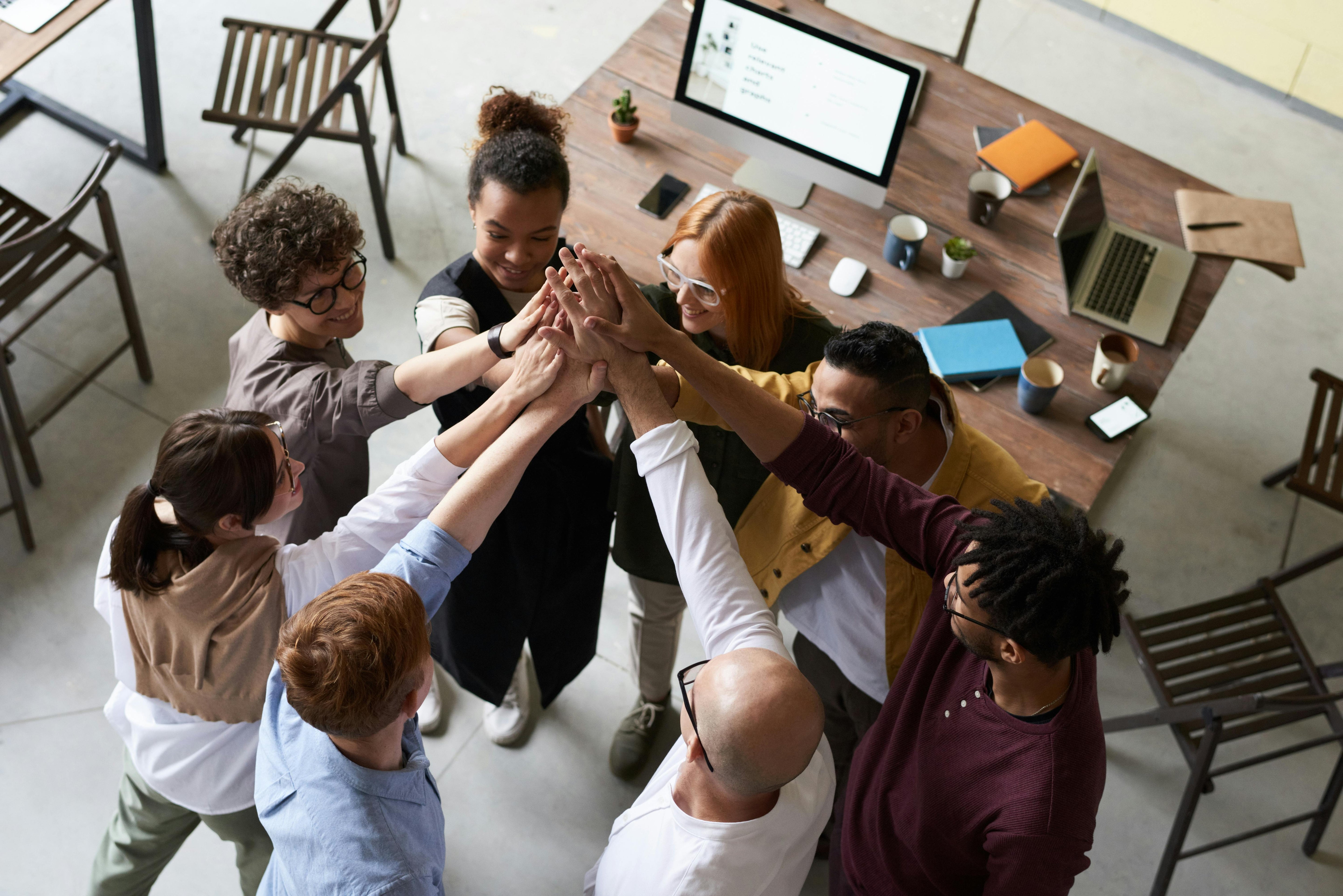 A diverse team standing in a circle with hands joined in the center, symbolizing teamwork, unity, and collaboration in a modern office setting.