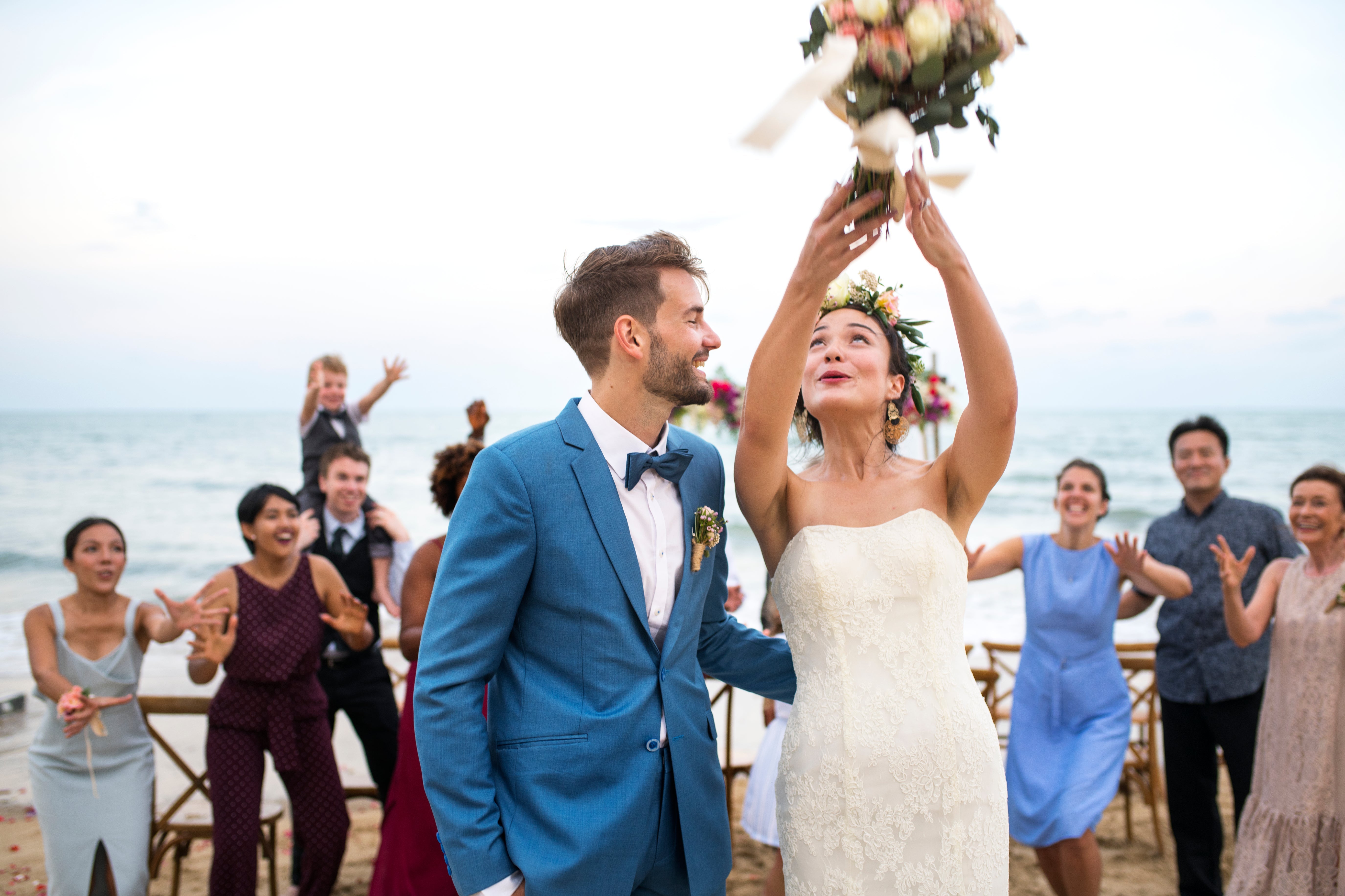 A joyful newly married couple celebrates their wedding on a beach, surrounded by excited family and friends, as the bride tosses her bouquet.