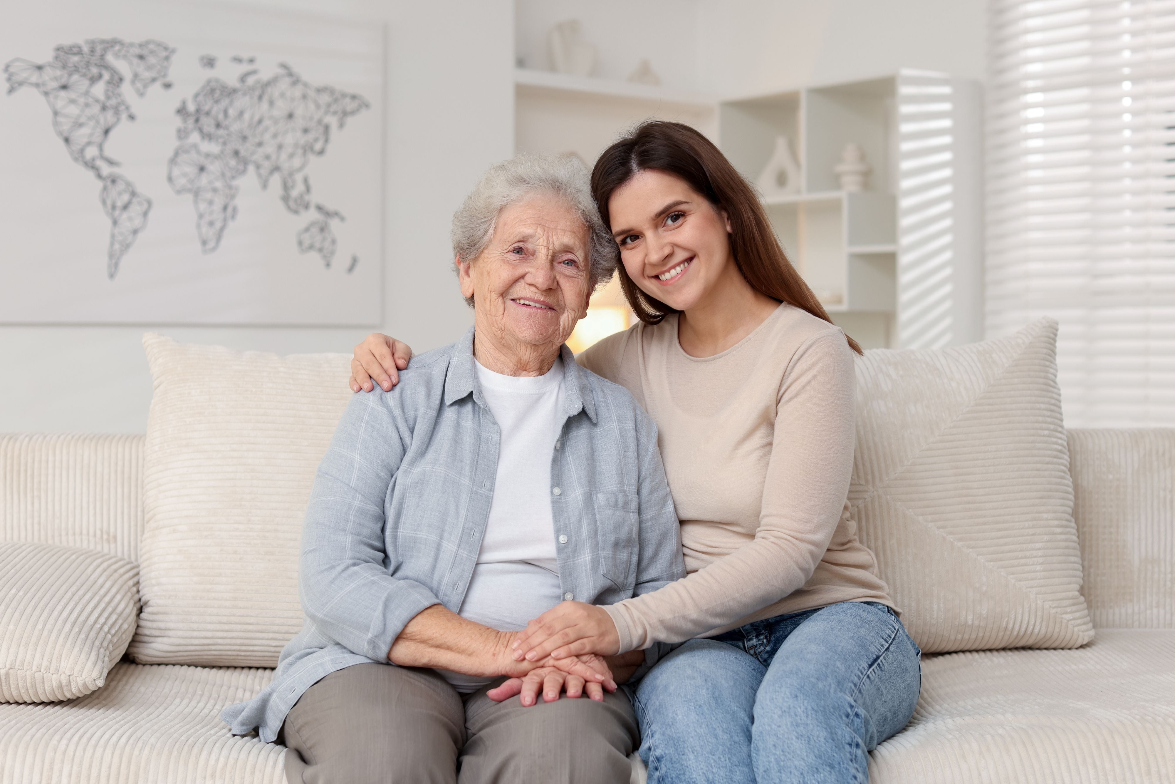 A smiling younger caregiver sits with her arm around an older parent on a light-colored sofa, illustrating warmth, support, and the importance of organized elder care.